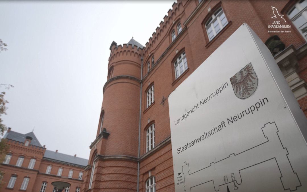 rotes Backsteingebäude im Hintergrund. Im Vordergrund ein silbernes Schild mit der Schrift Staatsanwaltschaft Neuruppin.